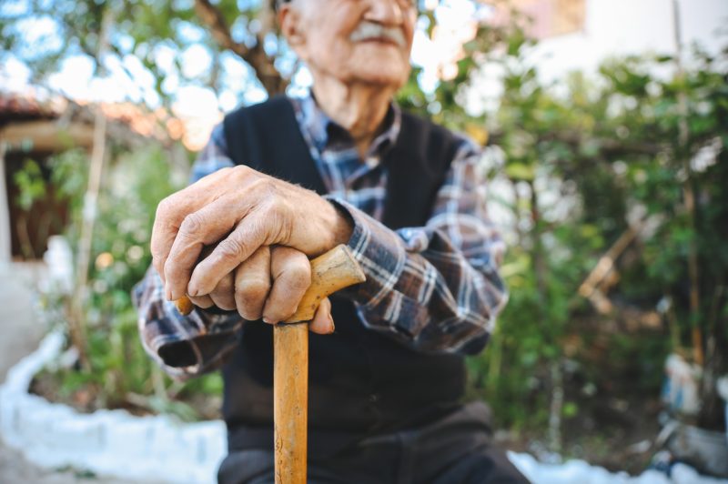 Old senior man with hands over the cane sitting in public park with happy expression. Elderly retiree with back pain using walking stick. Tree and flowers on background